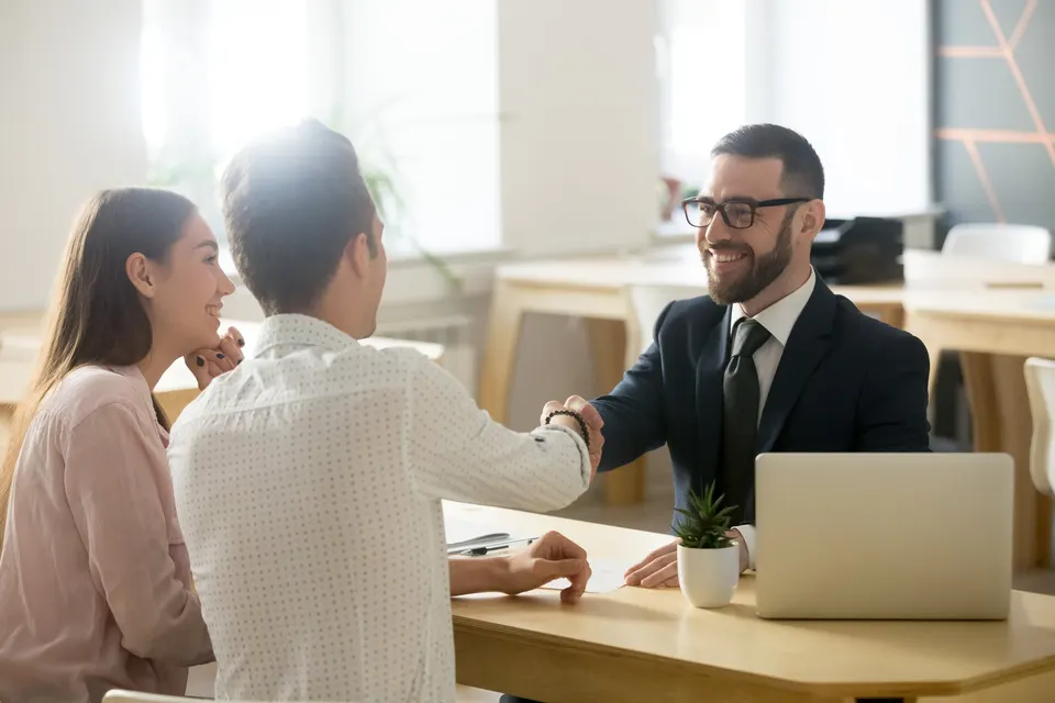 A banker in a suit shakes hands with a couple across a desk with a laptop.