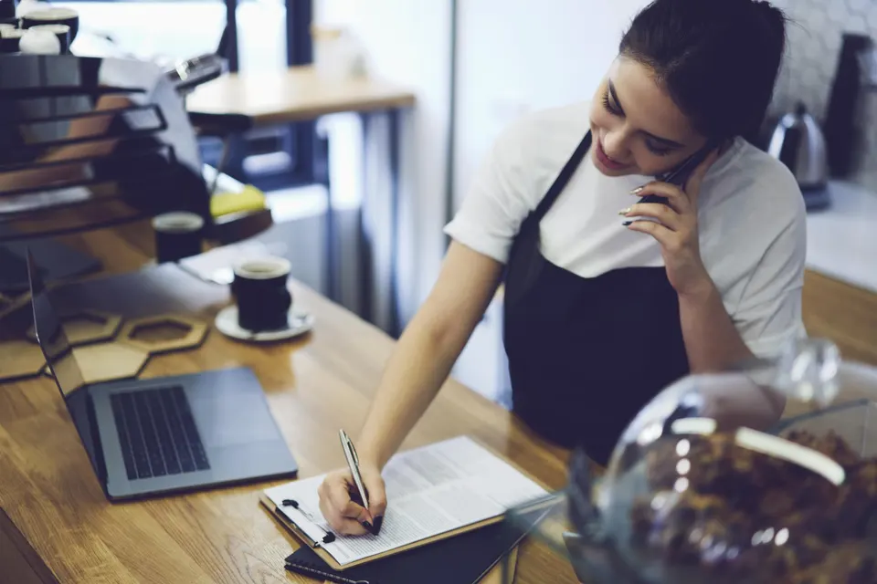 A café worker in an apron takes a phone order and writes it down at the counter beside a laptop.