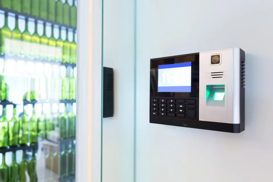 An access control terminal with a keypad, display and fingerprint reader mounted on a white wall beside a glass door.