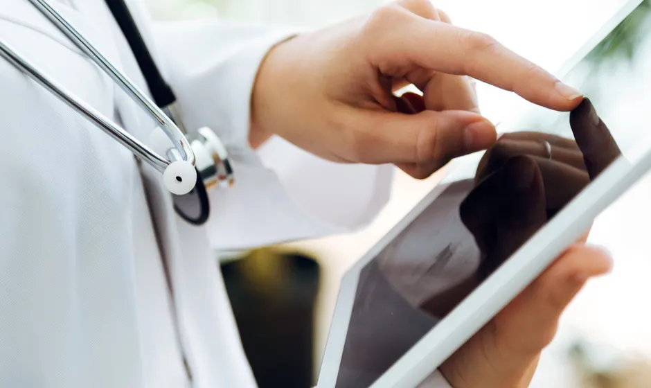 A clinician in a white coat with a stethoscope taps a tablet held in another person's hands.