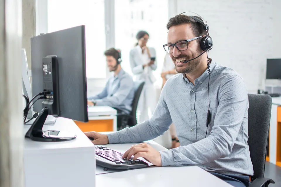 A smiling support agent in a headset works at a computer in a bright office.