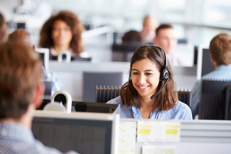A smiling agent wearing a headset works at a computer in a busy open-plan office.