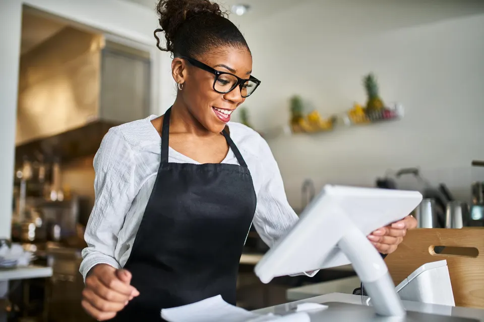 A smiling cashier in a black apron uses a countertop touchscreen POS terminal.