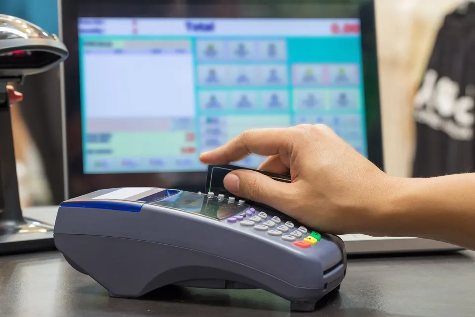 A card is swiped through a countertop payment terminal in front of a POS touchscreen.
