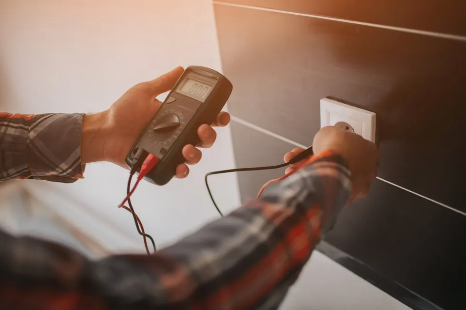 An electrician tests a wall outlet with the probes of a digital multimeter.