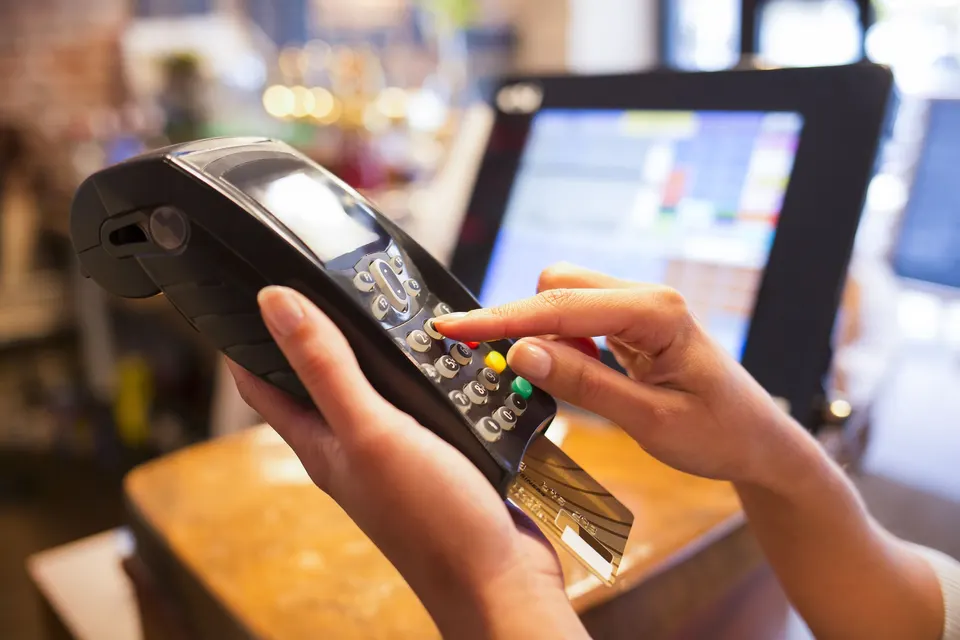 Hands holding a handheld card terminal and keying in a PIN at a store checkout, with a POS screen behind.