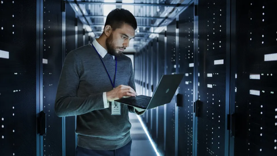 A technician works on a laptop in an aisle of server racks in a data center.