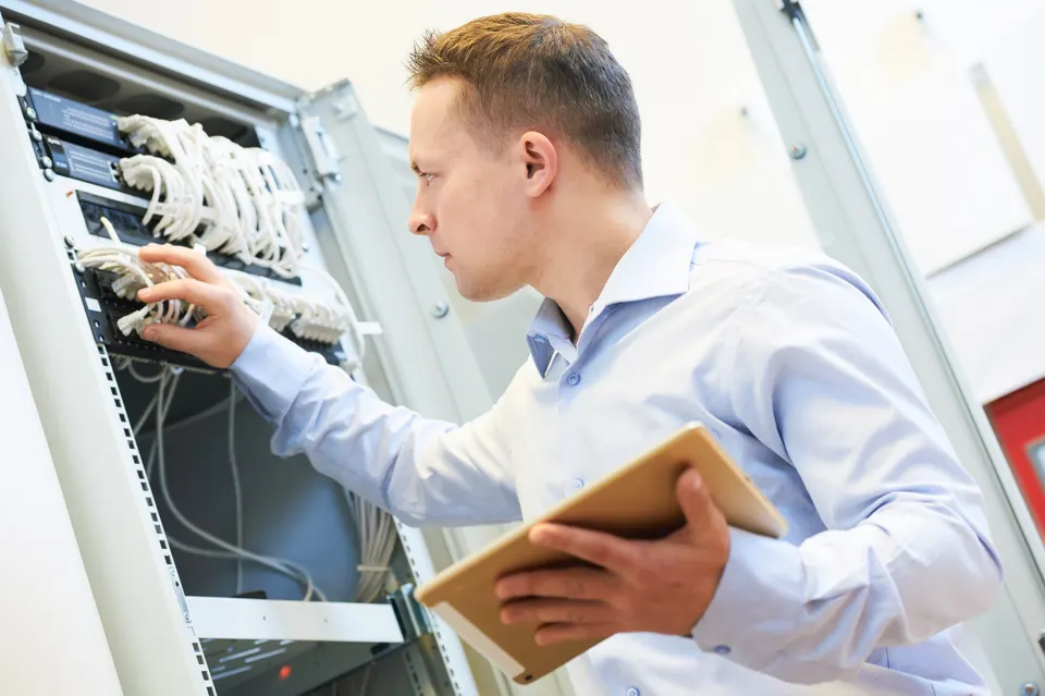 A technician holding a tablet checks the cabling inside an open network rack.